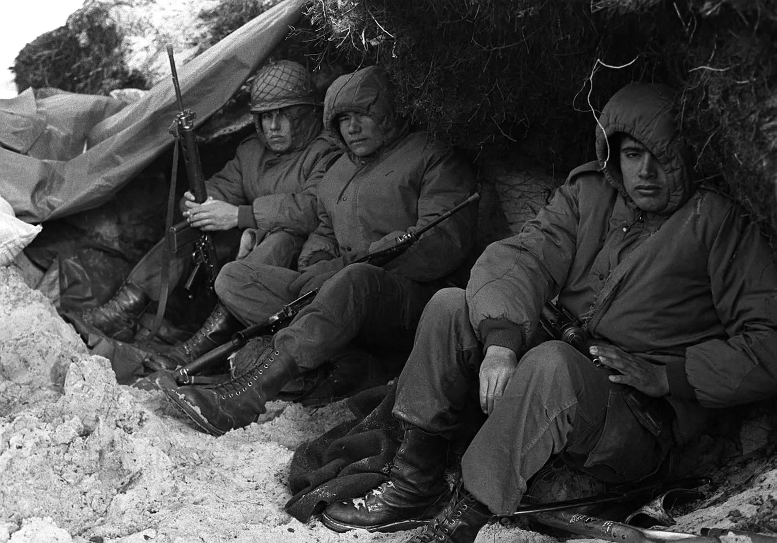 Tres soldados argentinos en una trinchera cubierta con lona, en blanco y negro: parkas, fusiles FAL y rostros de cansancio, Malvinas 1982.